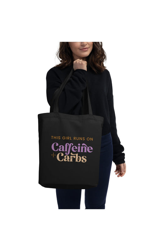 Woman holding a black cotton tote bag that says "This girl runs on caffeine + carbs"