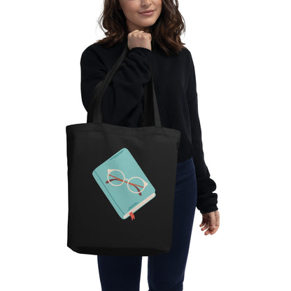 Woman hold black cotton tote bag with a cute illustration of a teal book with glasses on top