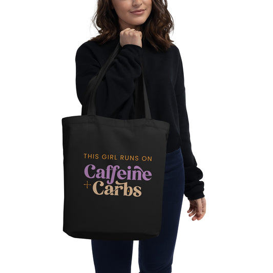 Woman holding a black cotton tote bag that says "This girl runs on caffeine + carbs"