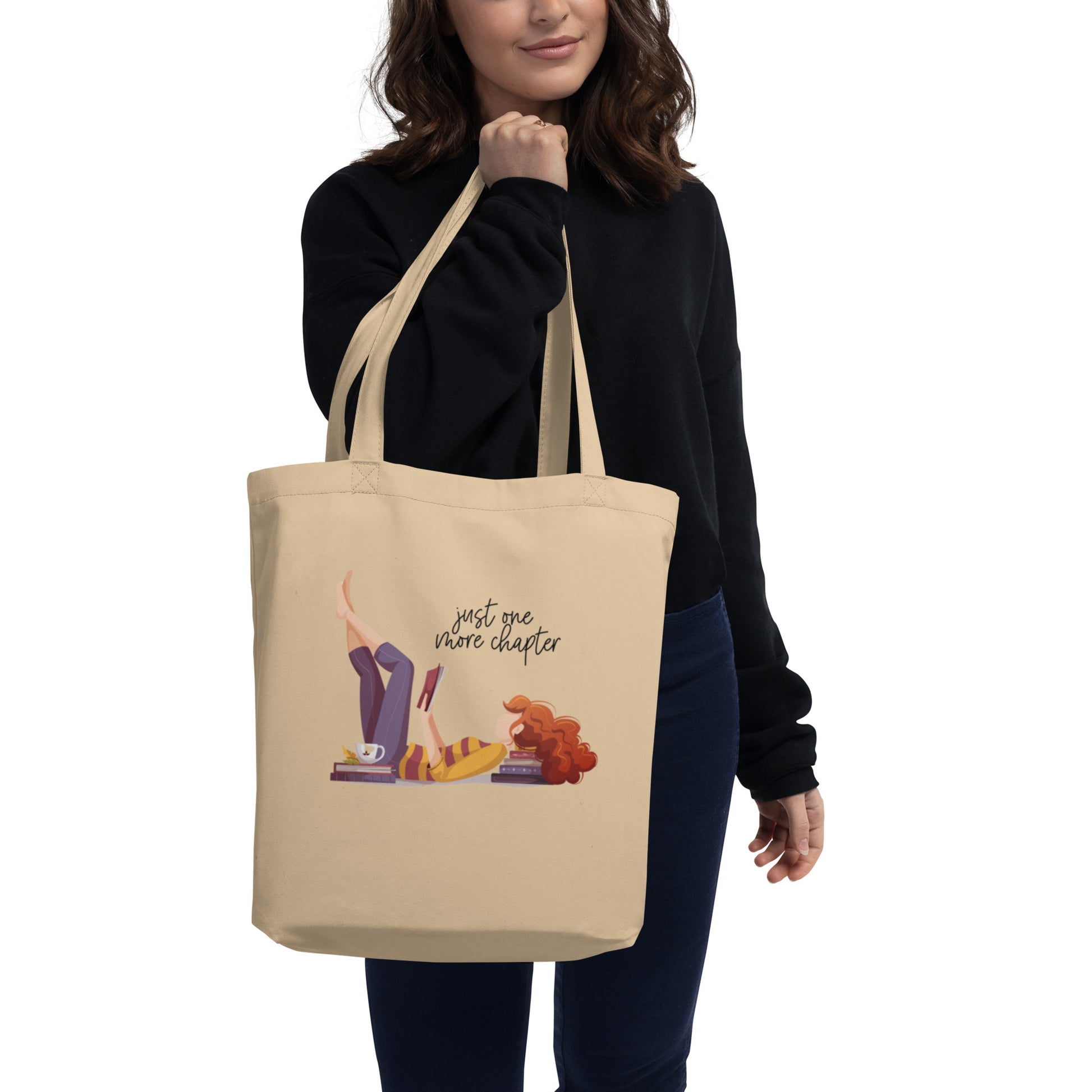 Woman holding tan tote bag that features artwork of a woman laying on the floor using a stack of books as a pillow. She's reading a book and the words say just one more chapter
