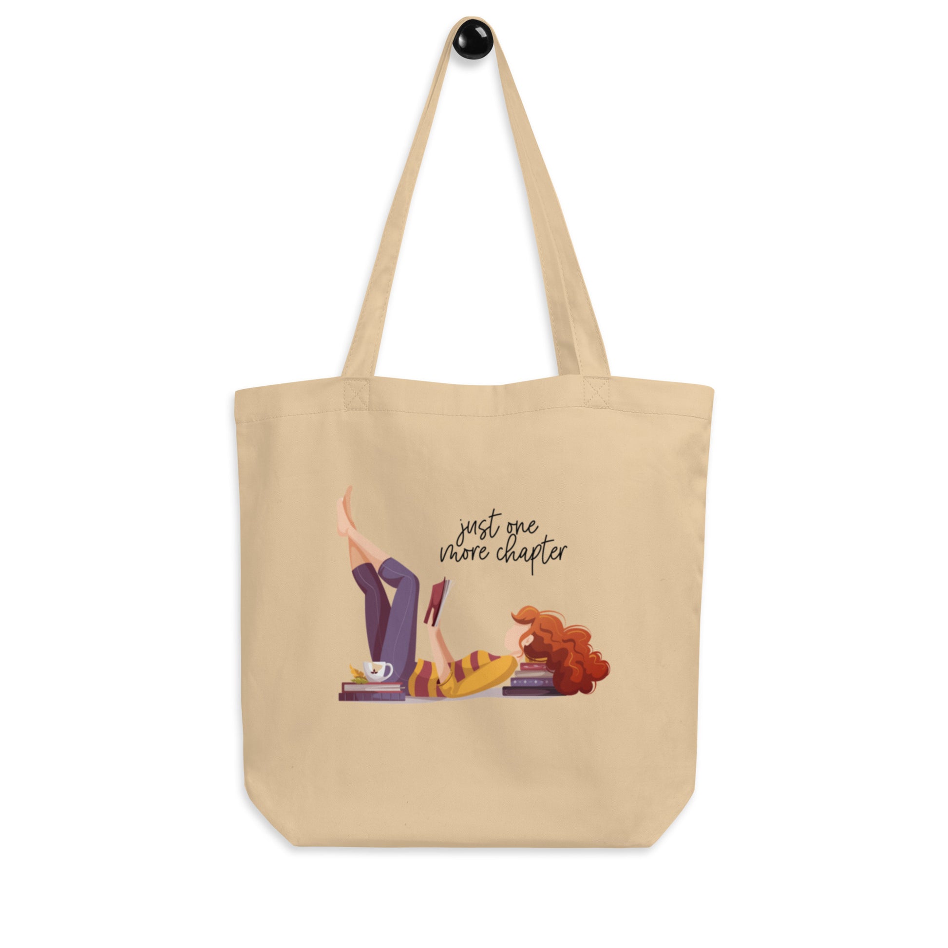 tan tote bag that features artwork of a woman laying on the floor using a stack of books as a pillow. She's reading a book and the words say just one more chapter